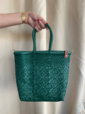 Green woven bag held by a hand against a beige curtain background